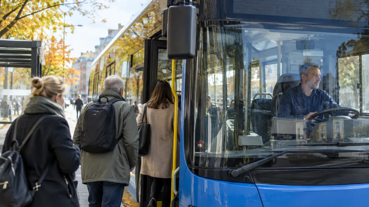 Bussförare vid ratten i en blå stadsbuss medan resenärer kliver på vid en hållplats en höstdag